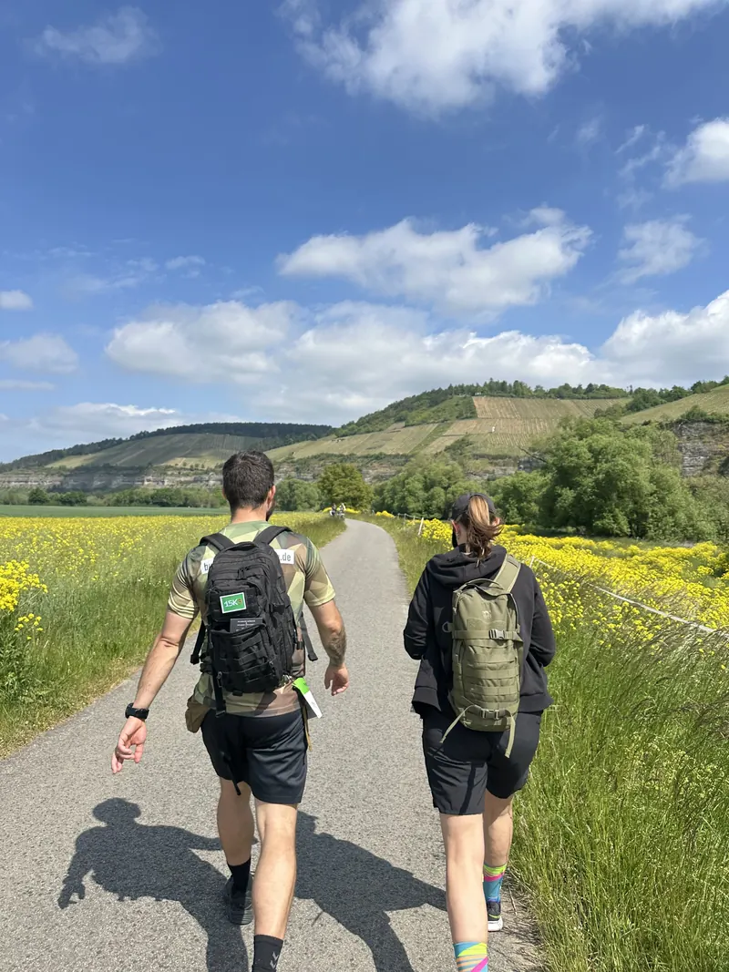 Zwei Teilnehmer mit Rucksack auf dem Weg am Main, links Rapsfeld